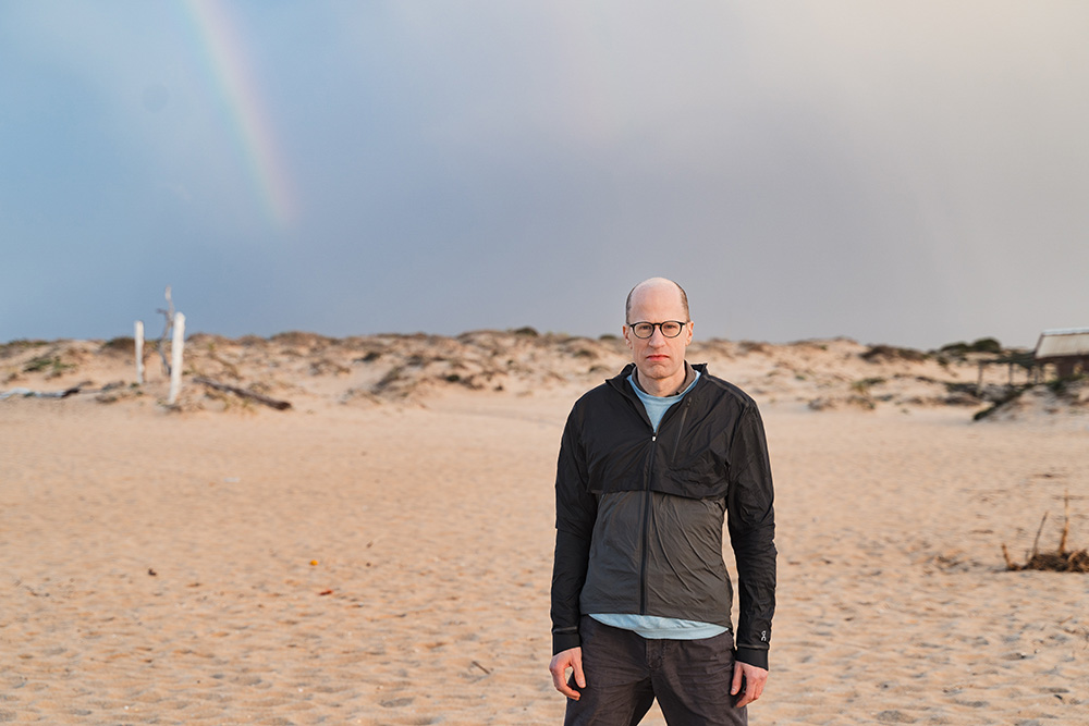 Outdoors portrait of Nick Bostrom in a black jacket at a beach with dunes in the background